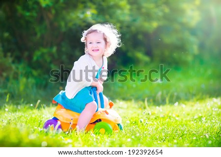 Happy laughing toddler girl in a blue dress having fun playing in the garden riding a yellow toy car enjoying sunny weather on a warm windy summer day