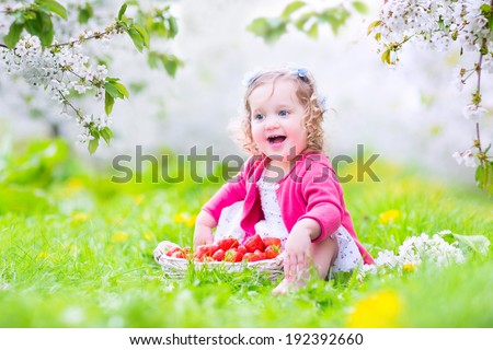 Cute happy toddler girl with curly hair and flower crown wearing red dress enjoying picnic in a beautiful blooming fruit garden with white blossoms on apple trees eating strawberry for healthy snack