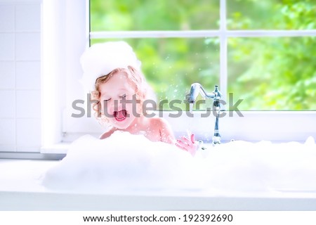 Funny little baby girl with wet curly hair taking a bath in a kitchen sink with lots of foam playing with water drops and splashes next to a big window with garden view
