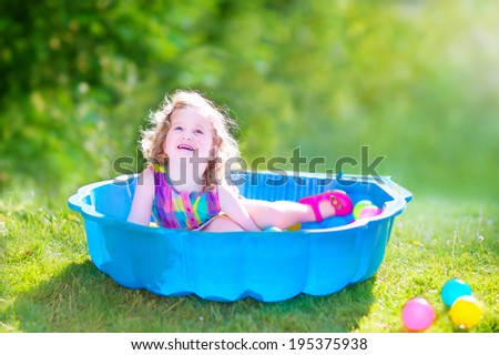 Happy cute toddler girl with curly hair wearing a pink colorful dress playing in a sand box with plastic toy balls in a sunny green garden on a hot summer day