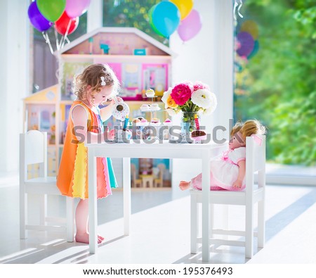 Adorable toddler girl with curly hair wearing a colorful dress on her birthday playing tea party with a doll, toy dishes, cup cakes and muffins in a sunny room with window