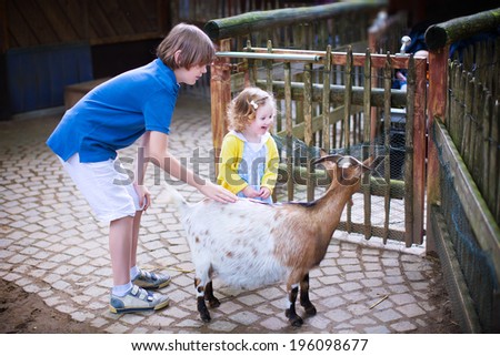 Happy laughing boy and his little toddler sister, adorable curly girl, playing together petting a goat laughing and having fun watching animals on a day trip to a modern city zoo