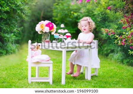 Adorable toddler girl with curly hair wearing a colorful dress on her birthday playing tea party with a teddy bear doll, toy dishes, cup cakes and muffins in a sunny summer garden