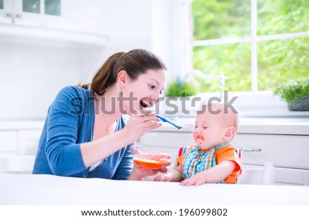 Young attractive mother feeding her cute baby son, giving him his first solid food, healthy vegetable pure from carrot with a plastic spoon sitting in a white sunny kitchen at a window at home