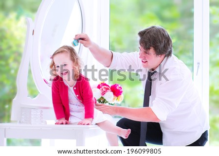 Young happy father in a business suit and tie brushing the hair of his daughter, cute little curly toddler girl, sitting on a white dresser with a beautiful round mirror in a white bedroom with window