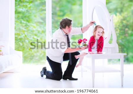 Young happy father in a business suit and tie brushing the hair of his daughter, cute little curly toddler girl, sitting on a white dresser with a beautiful round mirror in a bedroom with a big window