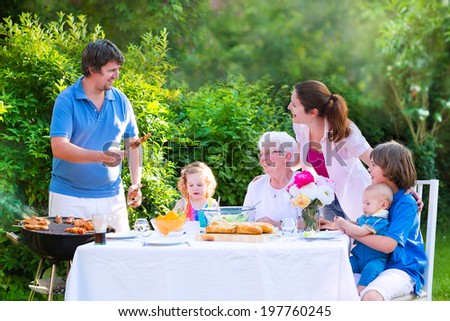 Happy big family - young mother and father with kids, teen age son, cute toddler daughter and a little baby, enjoying BBQ lunch with grandmother eating grilled meat in the garden with salad and bread
