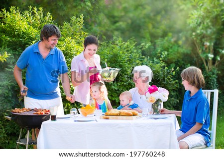 Happy big family - young mother and father with kids, teen age son, cute toddler daughter and a little baby, enjoying bbq lunch with grandmother eating grilled meat in the garden with salad and bread
