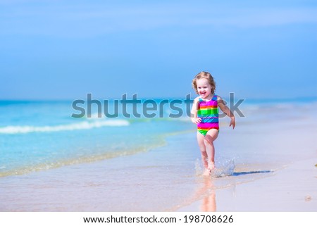 Happy laughing little girl in colorful rainbow bathing suit running and playing on ocean coast in water splashes on beautiful tropical island beach with turquoise clear water having fun on vacation