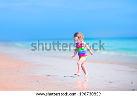 Happy laughing little girl in colorful rainbow bathing suit running and playing on ocean coast in water splashes on beautiful tropical island beach with turquoise clear water having fun on vacation