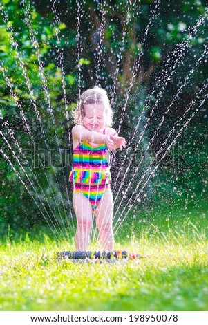 Funny laughing little girl in a colorful swimming suit running though garden sprinkler playing with water splashes having fun in the backyard on a sunny hot summer vacation day