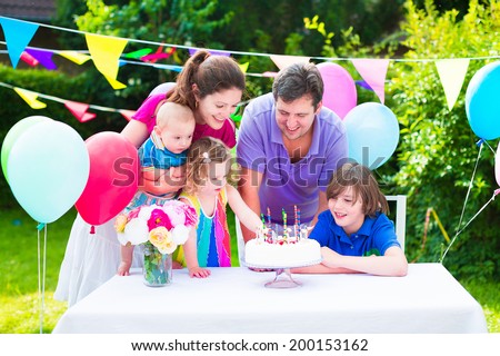 Happy big family with three kids - school age boy, toddler girl and a little baby enjoying birthday party with a cake blowing candles in a summer garden decorated with balloons and banners