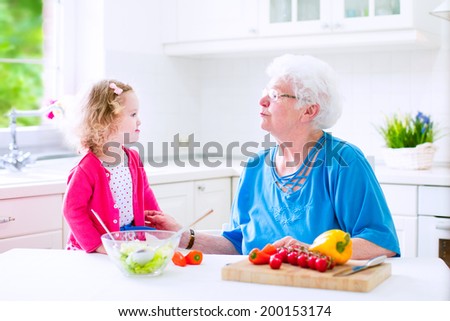 Beautiful senior lady, happy loving grandmother making healthy salad for lunch with her granddaughter, cute curly little girl in a white sunny kitchen with window, cutting pepper and tomato