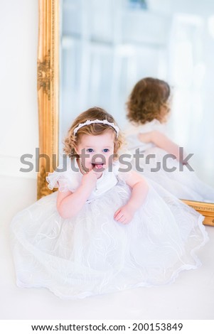 Sweet curly little girl with big beautiful eyes wearing a white bridesmaid dress sitting at a big window playing princess in a sunny living room