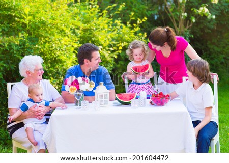 Happy big family - young mother and father with kids, teen age son, cute toddler daughter and a little baby, enjoying lunch with grandmother eating fruit, watermelon and strawberry in the garden