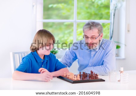 Happy loving grandfather enjoying a day with his grandson, laughing school age boy, playing chess in a white dining room with a window