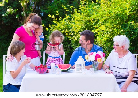 Happy big family - young mother and father with kids, teen age son, cute toddler daughter and a little baby, enjoying lunch with grandmother eating fruit, watermelon and strawberry in the garden
