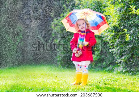 Funny cute curly toddler girl wearing red waterproof coat and yellow rubber boots holding colorful umbrella playing in the garden by rain and sun weather on a warm autumn or summer day