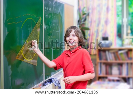 Happy smiling student boy enjoying his first day back to school at a mathematics lesson standing at a chalk board drawing for geometry study with triangle ruler on a big class room with window