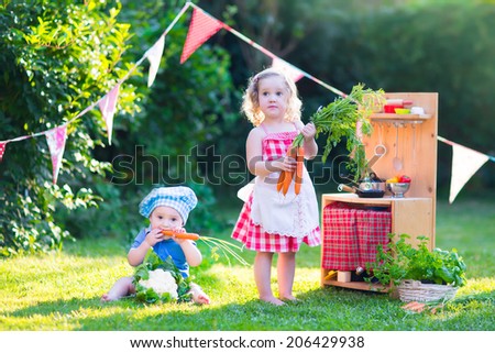 Funny curly little girl and adorable baby boy, cute brother and sister, playing together with a vintage wooden toy kitchen, table ware and fresh healthy vegetables in a sunny summer garden
