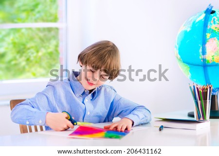 Happy smiling school boy, smart student, doing homework cutting paper, writing, drawing and reading a book at a white desk with a globe next to a window, back to school concept