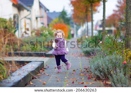 Little happy toddler girl in a warm purple jacket and boots playing in a beautiful city street with golden fall garden trees on a warm nice autumn day