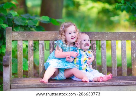 Adorable kids, little curly girl and a cute baby boy, brother and sister, sitting together on a wooden bench in a garden, hugging and kissing, relaxing and having fun on a sunny summer day