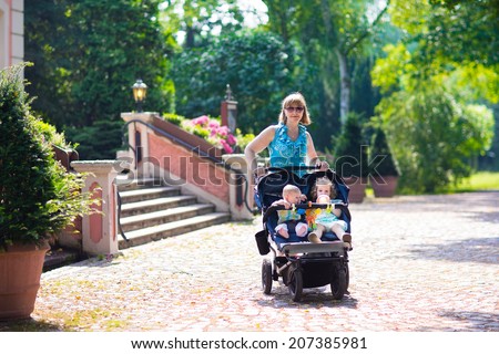 Young beautiful woman in a park with a double jogging stroller with two kids, baby boy and little toddler girl, brother and sister, walking fit and active on a hot summer day