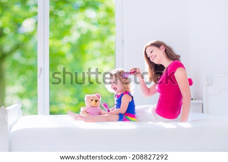 Happy family, beautiful young mother and her adorable little daughter, cute funny girl with curly hair brushing hair and playing with a teddy bear toy in a sunny white bedroom with window