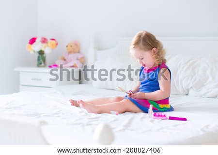 Cute little child, beautiful toddler girl with curly hair applying make up holding lipstick and mirror sitting on a white bed in a sunny bedroom playing with her toy teddy bear