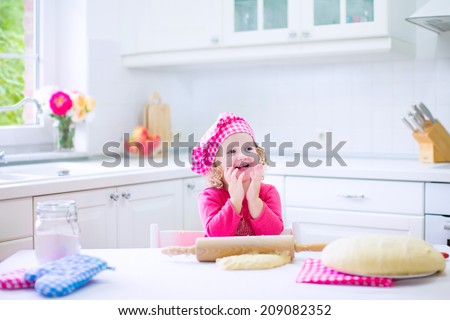 Adorable little child, funny curly toddler girl in a pink chef hat baking a pie rolling dough sitting in a white sunny kitchen with big window