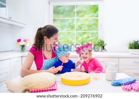 Young happy mother and her kids, adorable toddler girl and a little funny baby boy wearing pink and blue chef hats baking a pie together in a white sunny kitchen with big window