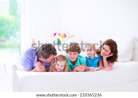 Happy big family, young parents with three kids, laughing boy, cute toddler girl and adorable little baby wearing colorful pajamas reading in bed in a white sunny bedroom at home