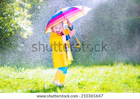 Funny cute curly toddler girl wearing yellow waterproof coat and boots holding colorful umbrella playing in the garden by rain and sun weather on a warm autumn or summer day