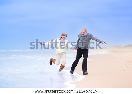 Happy middle aged couple, beautiful active woman and a laughing man running on a beach jumping away from waves on the North Sea, Scheveningen, Holland