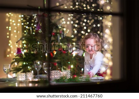 Cute curly toddler girl standing at a Christmas dinner table settling the dishes preparing to celebrate Xmas Eve, view through a window from outside into a decorated dining room with tree and lights