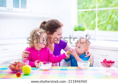Happy young family, mother with two children, adorable toddler girl and funny baby boy having healthy breakfast eating fruit and dairy, sitting in a white sunny kitchen with window