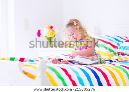 Cute curly little toddler girl reading a book sitting in a sunny bedroom on a wooden white bed with colorful rainbow bedding enjoying a nice weekend morning