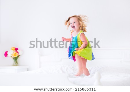 Cute little curly toddler girl in a colorful dress jumping on a big white bed laughing and having fun on a sunny weekend morning in a bedroom