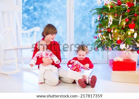 Three kids opening Christmas presents. Happy boy, little curly toddler girl and funny baby, brothers and sister, enjoying Xmas morning in a living room with decorated tree and window into snowy garden