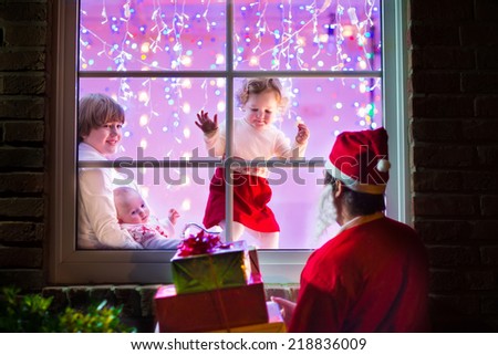 Three children, happy boy holding little baby and a cute toddler girl in a knitted winter dress watching their father in Santa costume standing outside of the house with presents, view through window