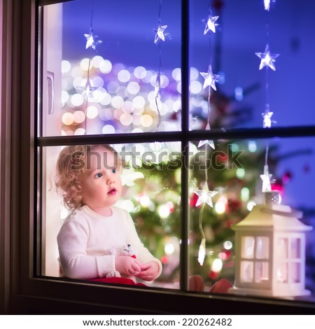 Cute curly toddler girl sitting with a toy bear at home during Christmas time, preparing to celebrate Xmas Eve, view through a window from outside into a decorated dining room with tree and lights