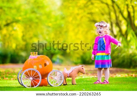 Cute curly little girl playing Cinderella fairy tale holding a magic wand next to a pumpkin carriage having fun in an autumn park at Halloween