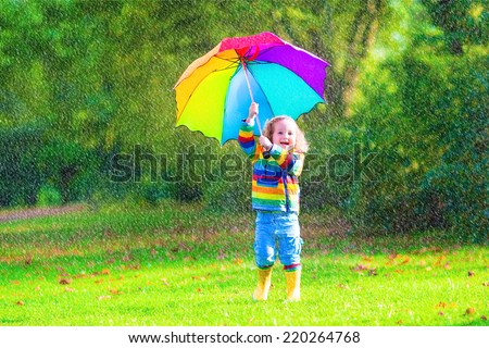 Funny cute curly toddler girl wearing yellow waterproof coat and boots holding colorful umbrella playing in the garden by rain and sun weather on a warm autumn or summer day
