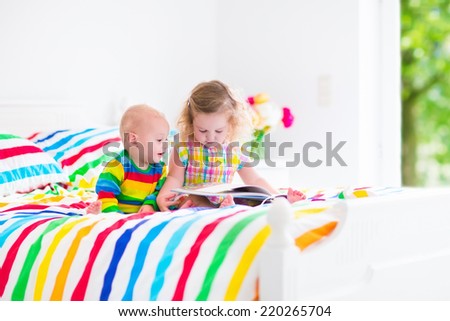 Two children, curly little toddler girl and funny baby boy, brother and sister, reading a book sitting in sunny bedroom on wooden white bed with colorful rainbow bedding enjoying nice weekend morning
