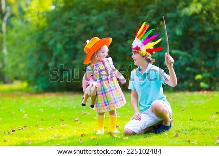 Two cute kids, laughing boy dressed as native American with colorful feather hat and funny curly toddler girl in yellow boots with a toy horse playing cowboy outdoors on a sunny summer day