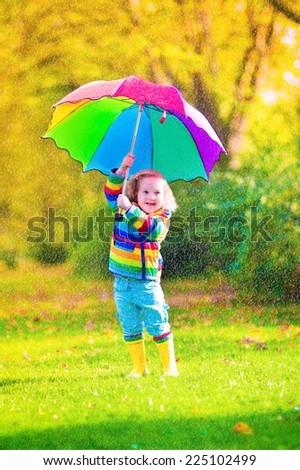 Cute curly toddler girl in yellow waterproof coat and boots holding colorful umbrella playing in the garden by rain and sun weather on a warm autumn or summer day