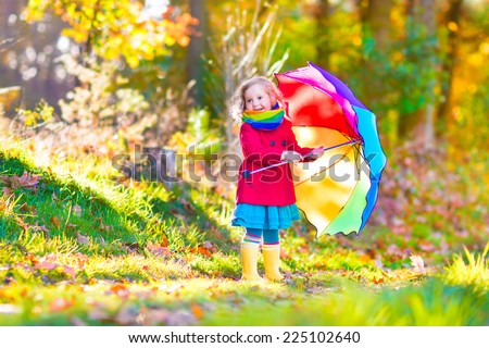 Cute curly little girl wearing a warm red coat and rain boots playing with colorful umbrella enjoying a sunny autumn day in a beautiful fall park