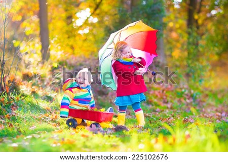 Cute little children, adorable toddler girl and a funny baby boy, brother and sister, playing in a sunny autumn park with a wheel barrow and colorful umbrella