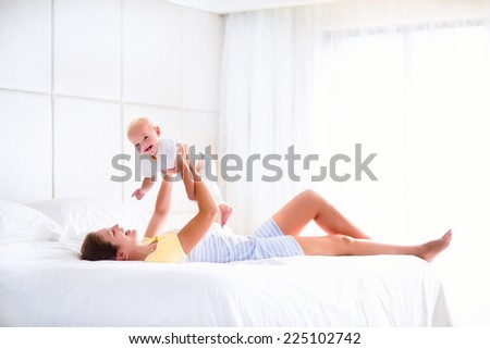 Young happy mother holding her adorable baby, cute blond curly boy, playing in a white bed enjoying a sunny morning in a modern home bedroom